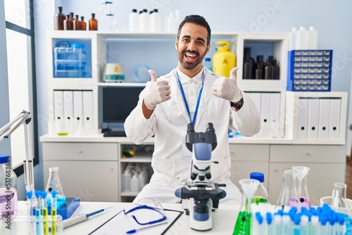 Young hispanic man with beard working at scientist laboratory success sign doing positive gesture with hand  thumbs up smiling and happy. cheerful expression and winner gesture.