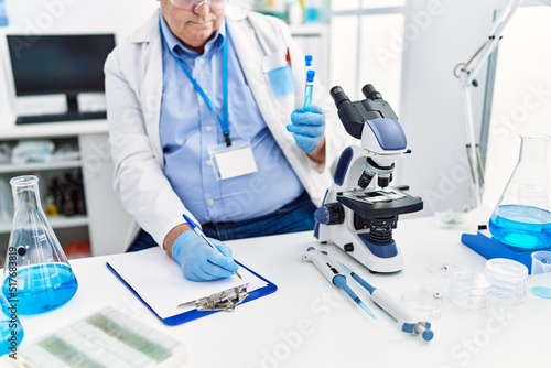 Middle age grey-haired man wearing scientist uniform holding test tube and write on document at laboratory