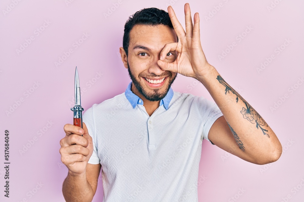 Hispanic man with beard holding pocket knife smiling happy doing ok sign with hand on eye looking through fingers Hispanic man with beard holding pocket knife smiling happy doing ok sign with hand on eye looking through fingers