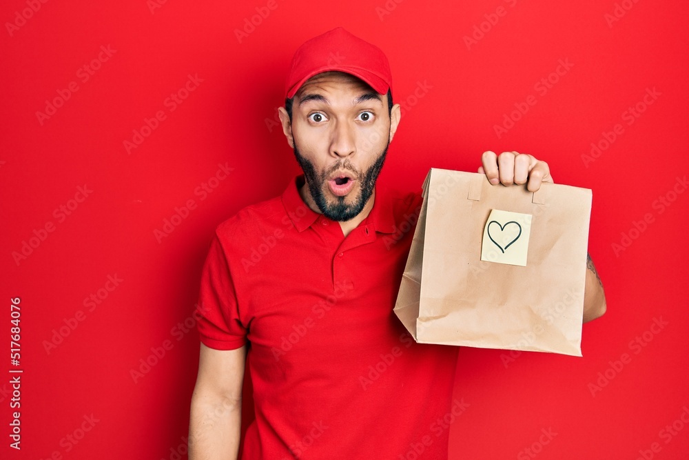 Hispanic man with beard holding take away paper bag with heart reminder scared and amazed with open mouth for surprise, disbelief face Hispanic man with beard holding take away paper bag with heart reminder scared and amazed with open mouth for surprise, disbelief face