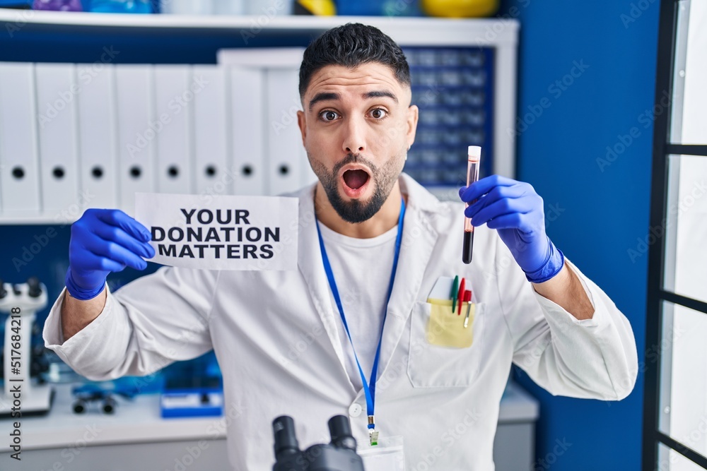 Young handsome man working at scientist laboratory holding blood sample afraid and shocked with surprise and amazed expression, fear and excited face. Young handsome man working at scientist laboratory holding blood sample afraid and shocked with surprise and amazed expression, fear and excited face.