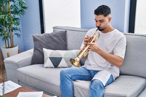Canvas Print Young arab man musician playing trumpet sitting on sofa at home