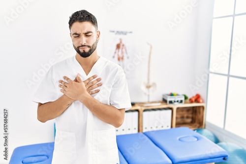 Young handsome man with beard working at pain recovery clinic smiling with hands on chest with closed eyes and grateful gesture on face. health concept.
