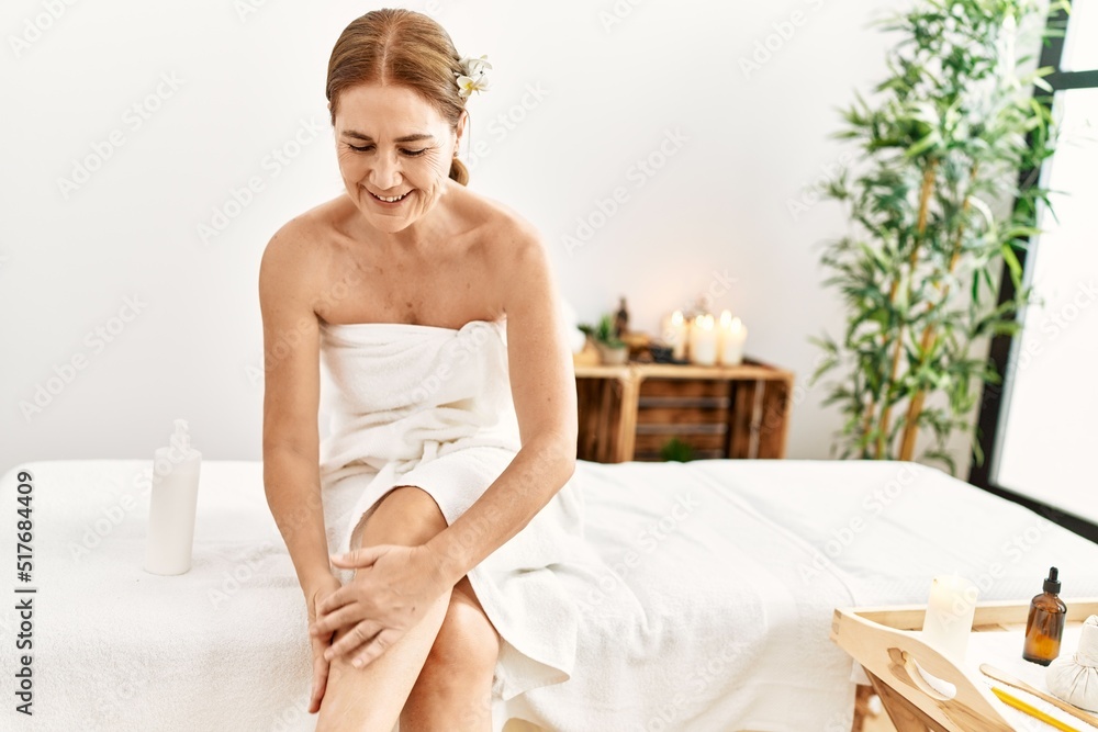 Middle age caucasian woman wearing towel massaging legs himself with cream at beauty center Middle age caucasian woman wearing towel massaging legs himself with cream at beauty center