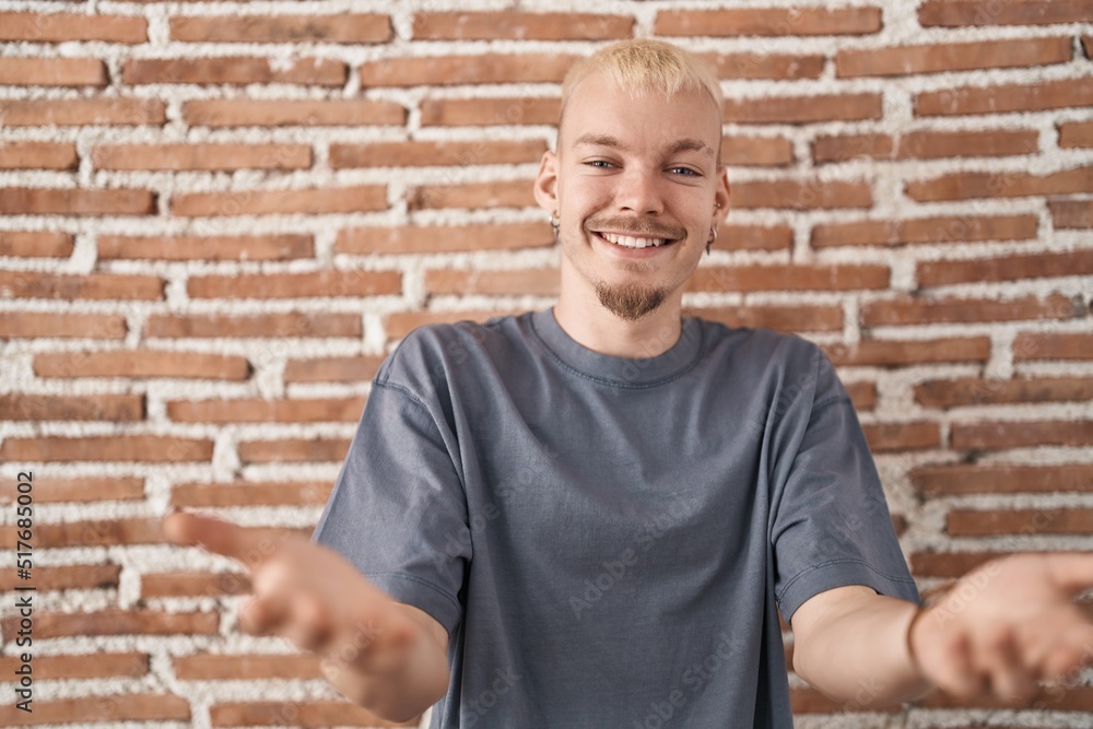 Young caucasian man standing over bricks wall smiling cheerful offering hands giving assistance and acceptance. Young caucasian man standing over bricks wall smiling cheerful offering hands giving assistance and acceptance.