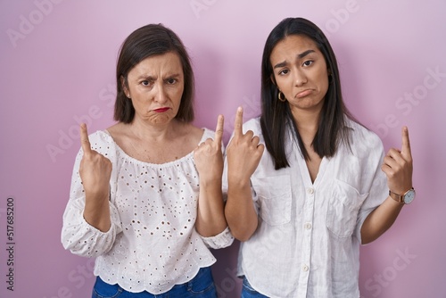 Hispanic mother and daughter together pointing up looking sad and upset, indicating direction with fingers, unhappy and depressed.