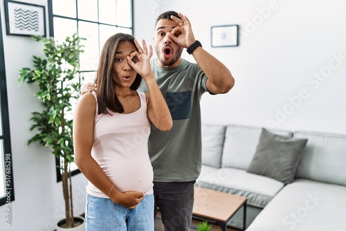 Young interracial couple expecting a baby, touching pregnant belly doing ok gesture shocked with surprised face, eye looking through fingers. unbelieving expression.