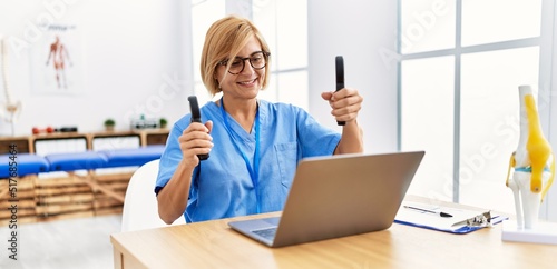 Fotografi Middle age blonde woman wearing physio therapy uniform doing tele rehab using ha