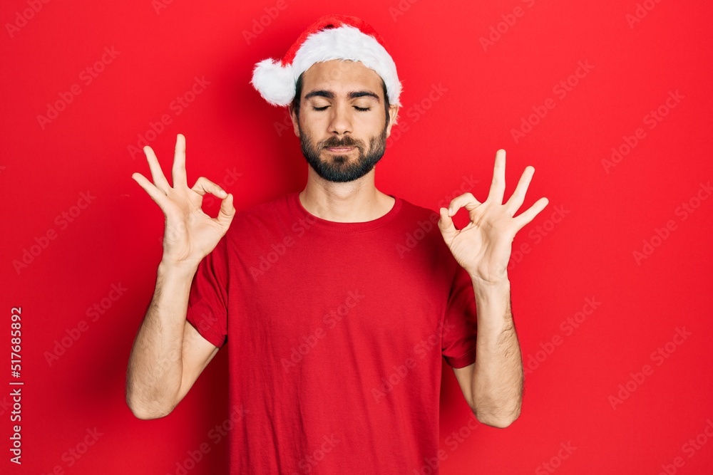 Young hispanic man wearing christmas hat relax and smiling with eyes closed doing meditation gesture with fingers. yoga concept. Young hispanic man wearing christmas hat relax and smiling with eyes closed doing meditation gesture with fingers. yoga concept.