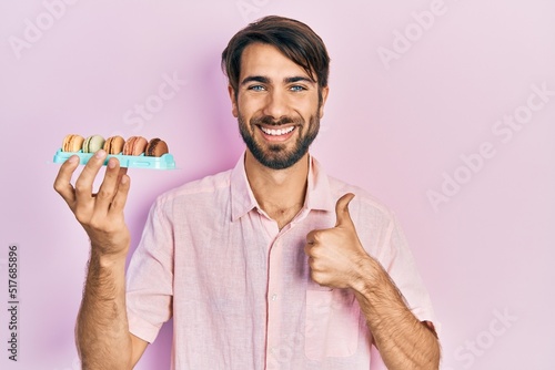 Young hispanic man holding delicious macrons pastries smiling happy and positive, thumb up doing excellent and approval sign photo