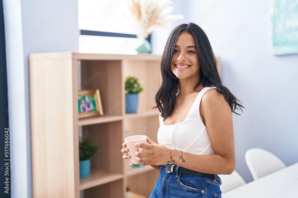 Young hispanic woman drinking coffee sitting on desk at home
