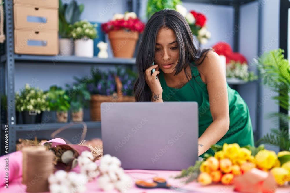 Young hispanic woman florist using laptop talking on smartphone at florist shop Young hispanic woman florist using laptop talking on smartphone at florist shop