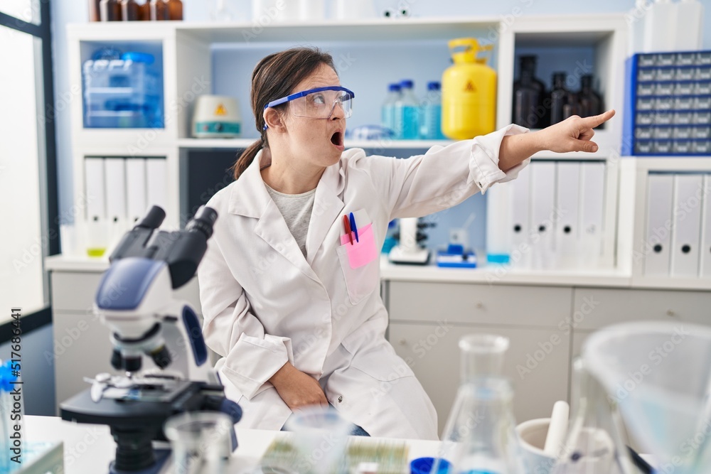 Hispanic girl with down syndrome working at scientist laboratory pointing with finger surprised ahead, open mouth amazed expression, something on the front Hispanic girl with down syndrome working at scientist laboratory pointing with finger surprised ahead, open mouth amazed expression, something on the front