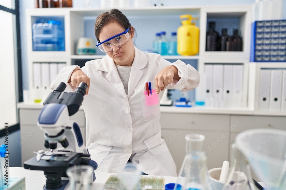 Hispanic girl with down syndrome working at scientist laboratory pointing down looking sad and upset, indicating direction with fingers, unhappy and depressed. Hispanic girl with down syndrome working at scientist laboratory pointing down looking sad and upset, indicating direction with fingers, unhappy and depressed.