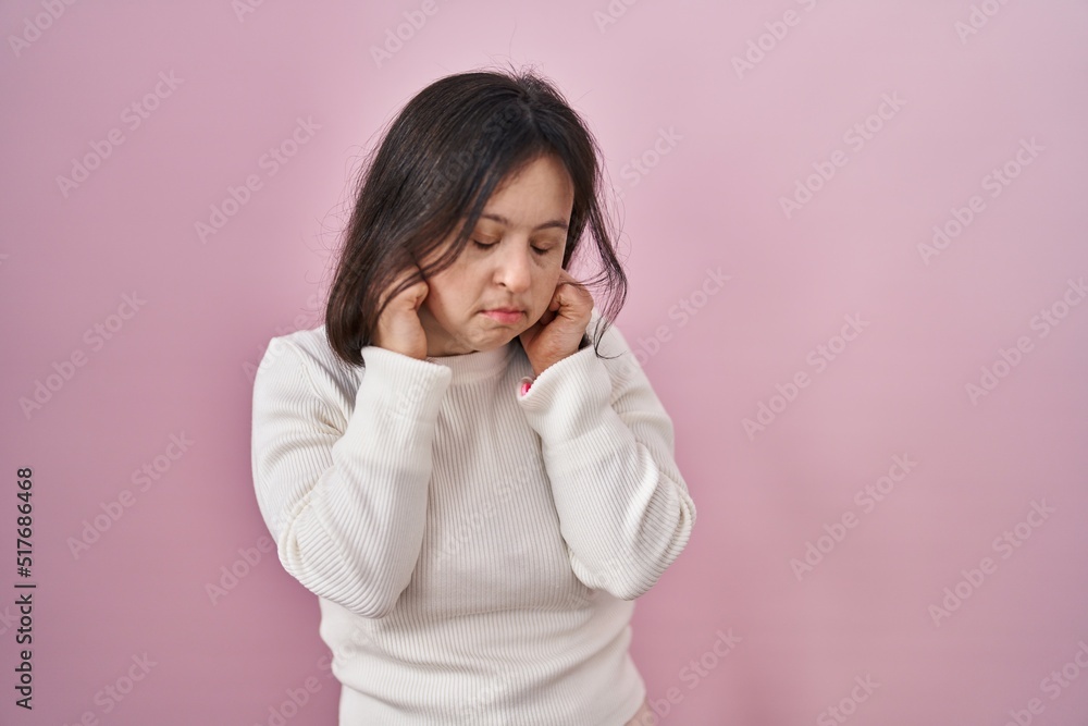 Woman with down syndrome standing over pink background covering ears with fingers with annoyed expression for the noise of loud music. deaf concept.
