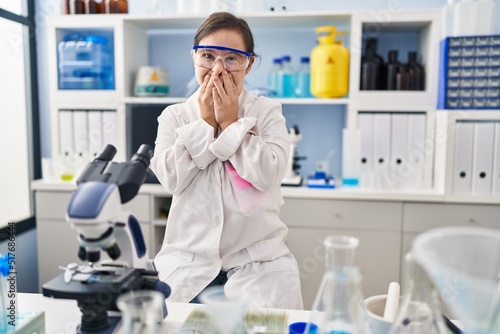 Hispanic girl with down syndrome working at scientist laboratory laughing and embarrassed giggle covering mouth with hands  gossip and scandal concept