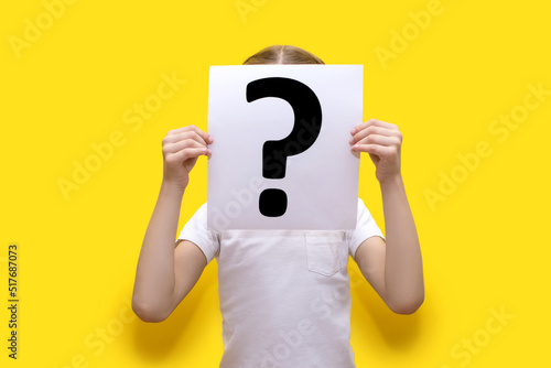 The child holds a sheet of paper with a question mark on a yellow background and covers his face. The concept of curiosity or indecision. Question mark on white paper in child's hands