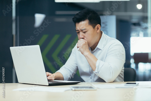 Sick man in office coughs and has runny nose, Asian businessman with cold works in office on laptop