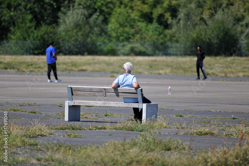 An elder man sits on a bench in the Tempelhof-Field in Berlin while he watches sportive people.