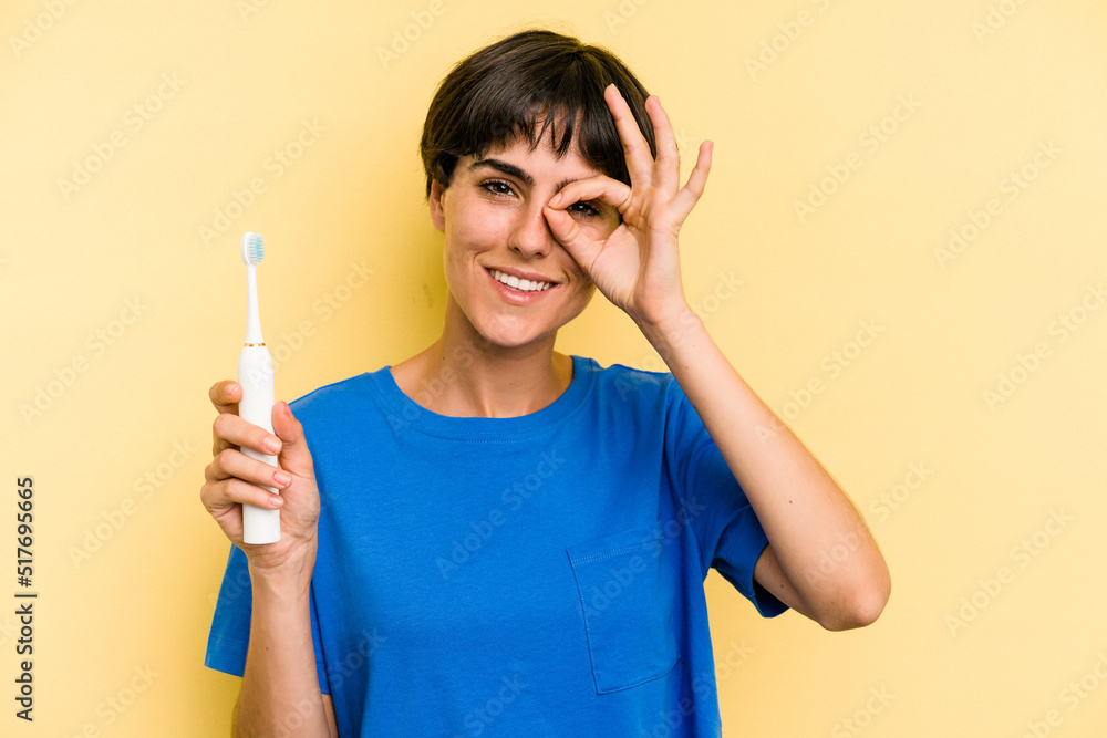 Young caucasian woman holding a electric toothbrush isolated on yellow background excited keeping ok gesture on eye. Young caucasian woman holding a electric toothbrush isolated on yellow background excited keeping ok gesture on eye.