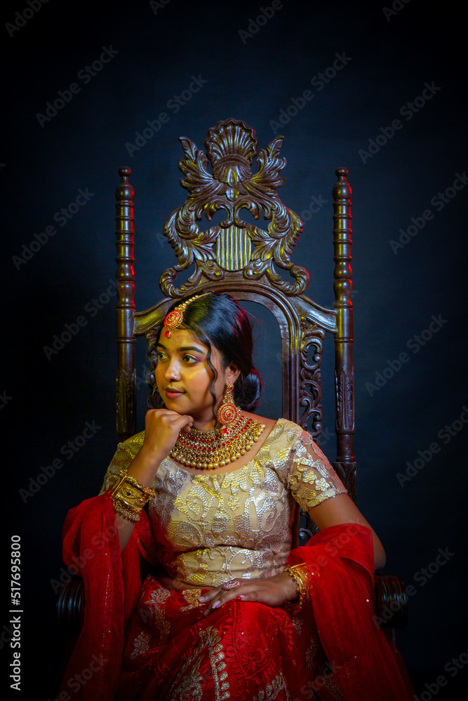 Princess lady in red traditional dress with gold jewellery . Indian Princess tradition costume golden jewellery set model sitting in a antique queen wooden chair with perfect makeup
