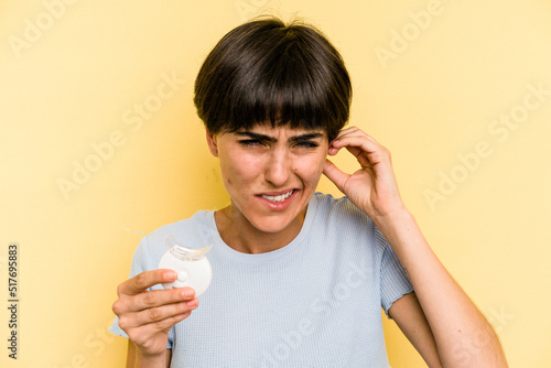 Young caucasian woman holding teeth whitener isolated on yellow background covering ears with hands.