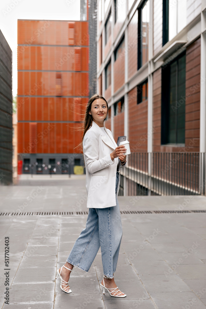 Full-lenght portrait of stylish attractive pretty woman with long hair wearing white jacket, jeans and heels is holding coffee and walking to office in business district