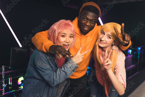 Multiracial young cheerful friends standing close together posing at camera in a dark gaming room with neon lights showing peace and thumbs up signs. High quality photo