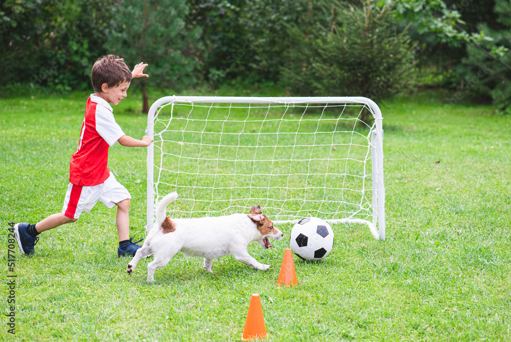Kid boy in red and white kit playing football (soccer) with his dog at training ground Kid boy in red and white kit playing football (soccer) with his dog at training ground
