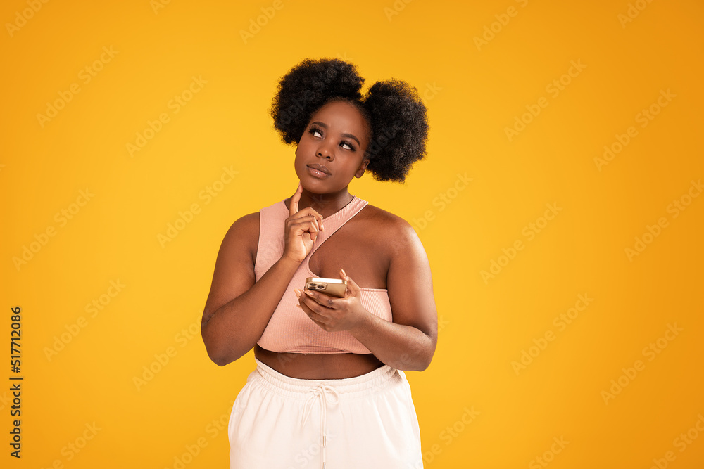 Young afro woman holding mobile phone and thinking about buying something online. Sale concept. Studio shot. Young afro woman holding mobile phone and thinking about buying something online. Sale concept. Studio shot.