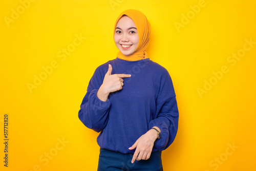 Cheerful young Asian Muslim woman dressed in casual sweater pointing at herself with proud isolated over yellow background © Sewupari Studio