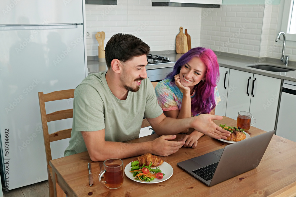 Hipster couple having a breakfast with croissant and salad, streaming a show. Young woman with colorful hair and bearded man at the kitchen watching memes on laptop. Close up, copy space, background Hipster couple having a breakfast with croissant and salad, streaming a show. Young woman with colorful hair and bearded man at the kitchen watching memes on laptop. Close up, copy space, background