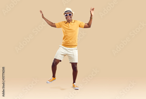 Cool cheerful african american man funny rejoicing and dancing isolated on beige background. Emotional dark-skinned man in stylish summer clothes having fun on vacation. Fun concept. Full length.