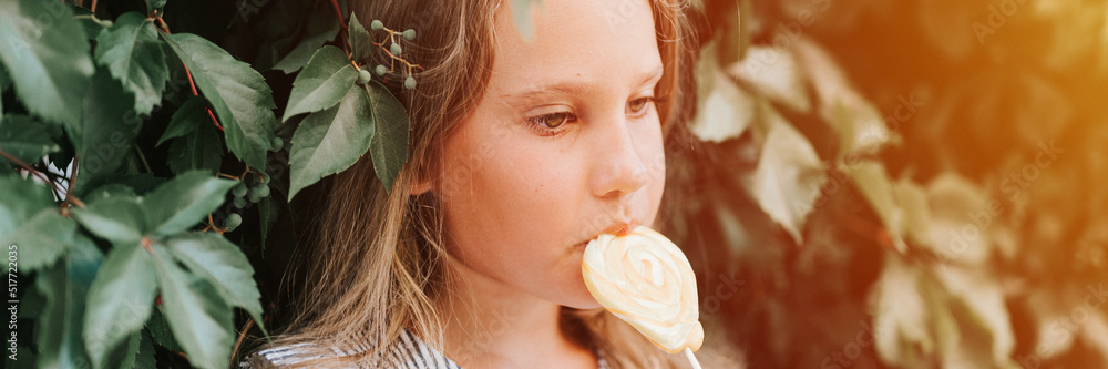 candid thoughtful little kid girl of eight years old holding lollipop and lick eating fruit sugar sweet candy on the background of hedge wall of green plants in summer vacation outdoor. banner. flare