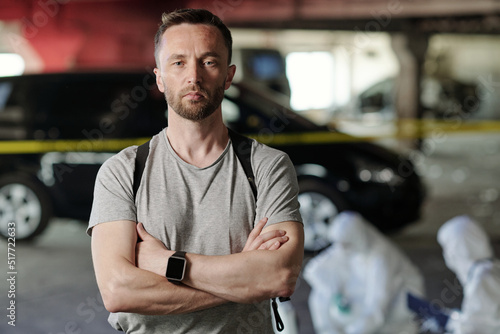 Young serious crime scene worker or detective inspector looking at camera with his arms crossed on chest standing against experts © pressmaster