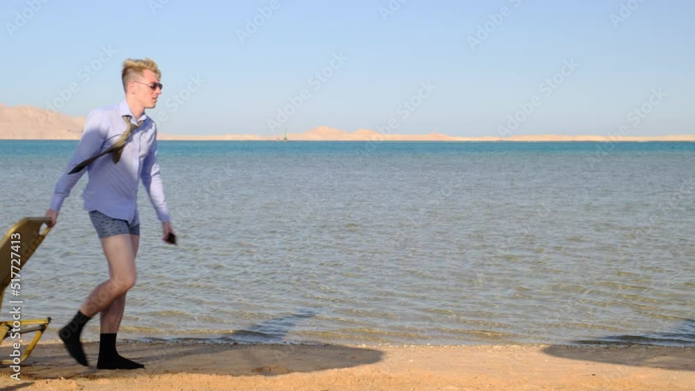 Funny businessman wearing shirt with tie and underwear with phone in his hand walking along beach against sea and drags sunbed behind him. Work during rest. Vacation. Slow motion Funny businessman wearing shirt with tie and underwear with phone in his hand walking along beach against sea and drags sunbed behind him. Work during rest. Vacation. Slow motion