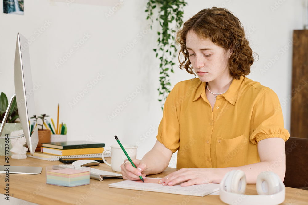 Young creative female freelancer with ruler and pencil drawing sketch in copybook while sitting by desk in front of computer monitor Young creative female freelancer with ruler and pencil drawing sketch in copybook while sitting by desk in front of computer monitor