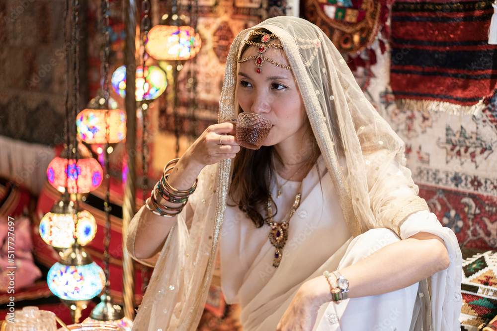 Asian woman wearing a necklace and a traditional Indian dress wrapped in a sari sits in a restaurant to drink tea.