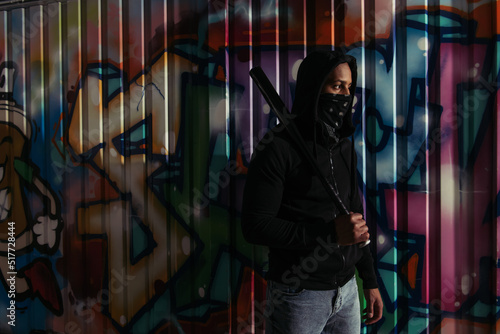 African american vandal with mask on face holding baseball bat near graffiti on urban street