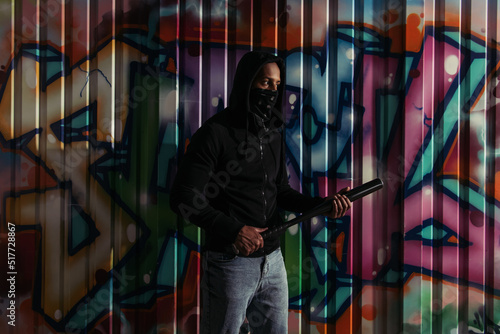 African american vandal in hoodie holding baseball bat near graffiti outdoors at night