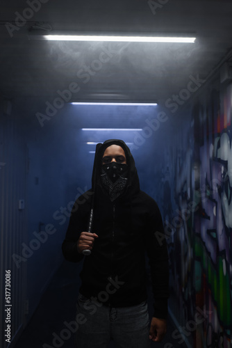 African american hooligan in mask holding baseball bat and looking at camera near lighting and graffiti