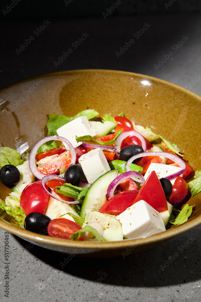 Classic greek salad on ceramic bowl in minimal style. Simple composition with greek salad on gray stone background with hard shadow. Dark concept menu. Vegetable salad on concrete table and shadow.