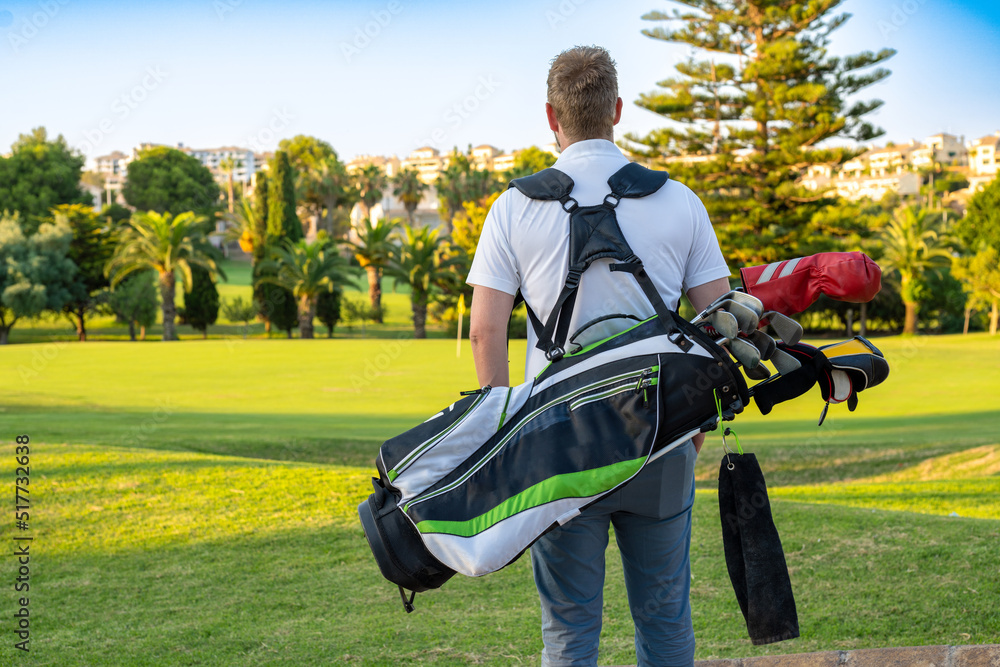 Back view of young handsome man at the course carrying a golf bag