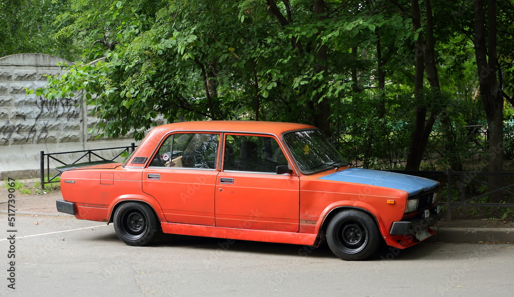 An old red Soviet car with a blue hood is parked next to the lawn, Podvoysky Street, St. Petersburg, Russia, July 2022
