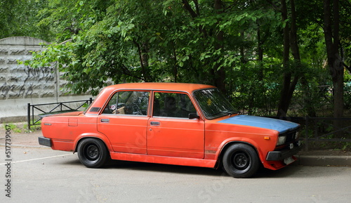 An old red Soviet car with a blue hood is parked next to the lawn  Podvoysky Street  St. Petersburg  Russia  July 2022