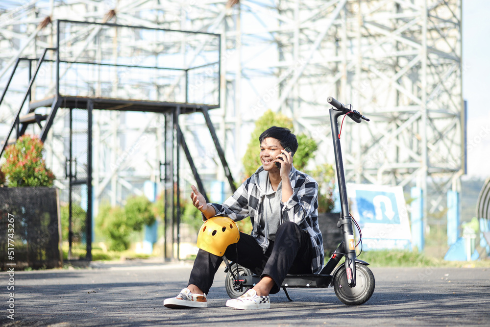 Asian man talking on the phone while sitting on electric scooter at the park street. Modern and ecological transportation concept. Asian man talking on the phone while sitting on electric scooter at the park street. Modern and ecological transportation concept.