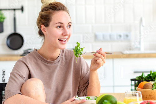Beautiful young woman eating fresh salad sitting at the kitchen table at home. Vegan food. Healthy lifestyle concept
