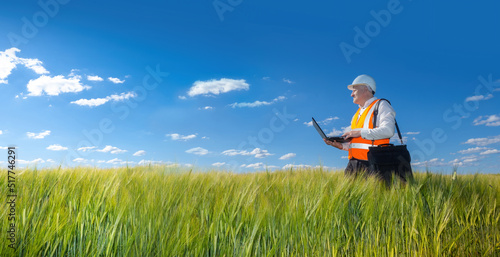 A man in a helmet and an orange vest walking along the field with grass. engineer builder examines the place of future construction. A man with a laptop stands in a field with grass.