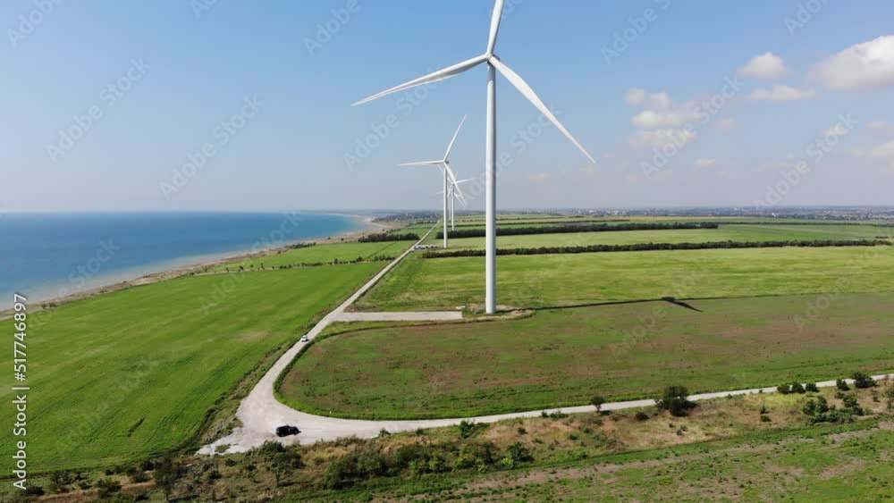 Aerial view 4K video. Wind power plant. Panoramic landscape with windmills in the green agriculture fields and the seashore. Renewable energy generation. The car rides along the road. Summer nature Aerial view 4K video. Wind power plant. Panoramic landscape with windmills in the green agriculture fields and the seashore. Renewable energy generation. The car rides along the road. Summer nature
