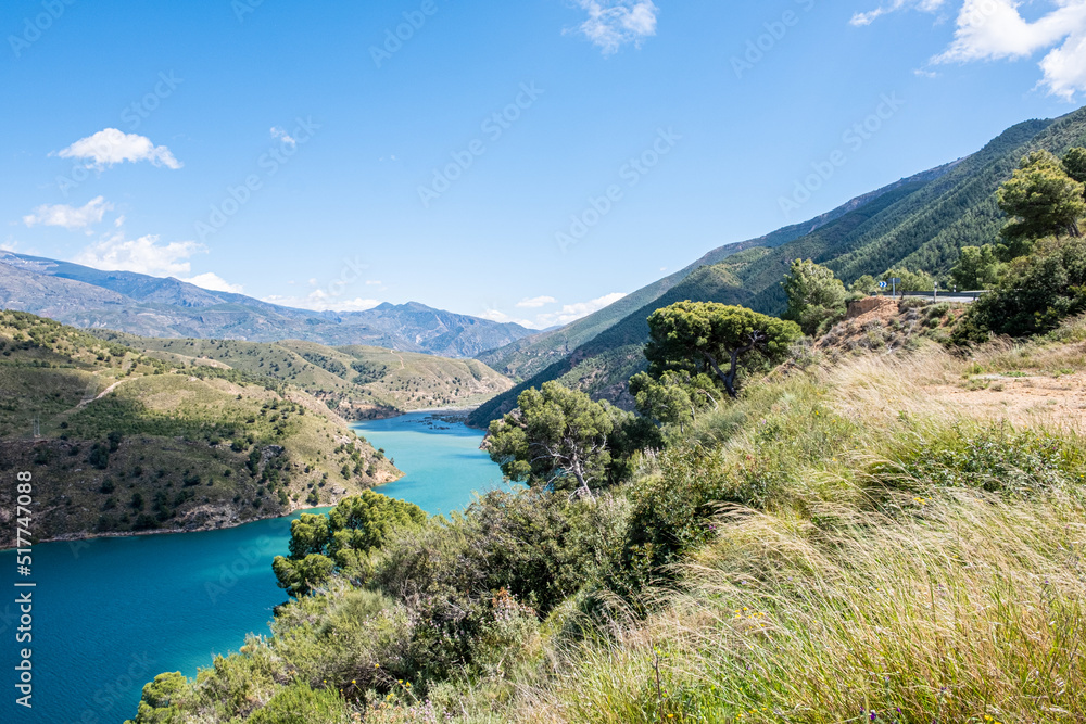 Breath-taking view from the top on green lake surrounded by picturesque mountain alley with trees and bushes in sunny summer day with blue sky and white clouds in background.